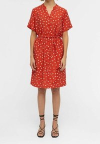 Rød polka dot kjole med V-udskæring, korte ærmer, snøret talje, knælang fald og let stof. Båret med sorte sandaler med remme.