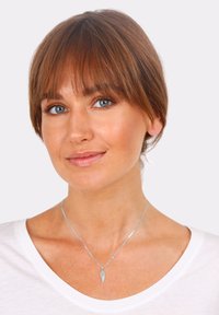 Collana d'argento con un pendente a forma di ala, indossata sopra una maglietta bianca. Il pendente presenta dettagli intricati e una finitura lucida.