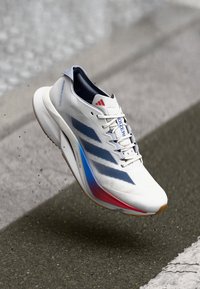 Hvid atletisk sko med blå og røde accenter, der har en mesh-overdel, polstret sål og tre-stribe design. Letvægts og moderne form.