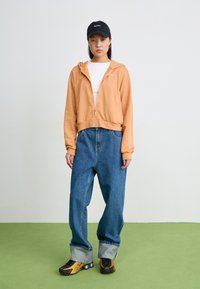 Oransje zip-up hettegenser, hvit t-skjorte, oversized blå jeans og svart caps med hvit tekst. Gule og svarte sneakers fullfører antrekket.