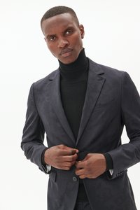 Chaqueta de traje de patrón oscuro con solapa notch, cierre de botón y dos bolsillos frontales, combinada con un suéter de cuello alto negro.
