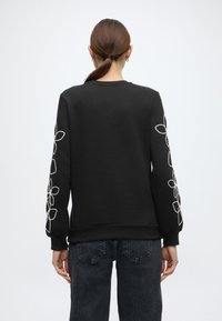 Μαύρο φούτερ με μακριά μανίκια, διακοσμημένο με λευκή κεντητική σε λουλουδάτα μοτίβα στο αρισ τερό μανίκι, στρογγυλή λαιμόκοψη και ριμπ μανσέτες και τελείωμα.