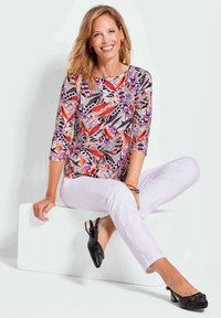 Bluse mit floralem Muster in Pink, Lila und Orange mit drei Viertelärmeln, kombiniert mit weißen Hosen und schwarzen Slingback-Schuhen mit Hardware-Akzenten.