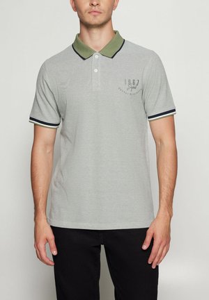 Mand iført grå polo med olivengrøn krave og ærmekanter, sorte bukser, stående mod en ensfarvet lys baggrund.