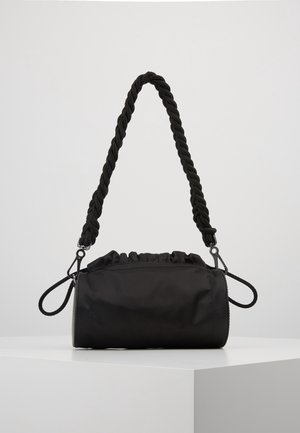 Schwarze zylindrische Handtasche mit gerafftem Oberteil und dickem, verdrehtem Seilschulterriemen, auf einem weißen Sockel vor hellem Hintergrund platziert.