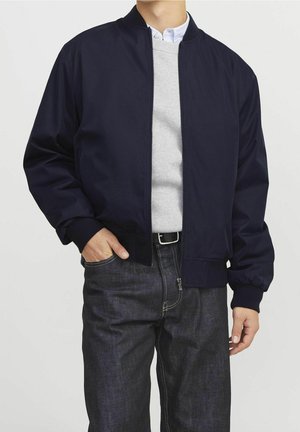 Homme portant une veste bomber bleu marine foncé sur un pull gris clair et une chemise boutonnée, associée à un jean foncé et une ceinture noire.