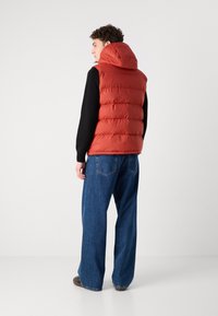 Rød puffervest med hette, med quiltede seksjoner og høy krage. Kombinert med løstsittende blå jeans.
