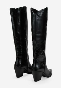 Botas negras hasta la rodilla con textura de cocodrilo, puntera afilada y tacón cuadrado. Presentan un cierre de cremallera en el lateral y bordes superiores con forma de ondas.