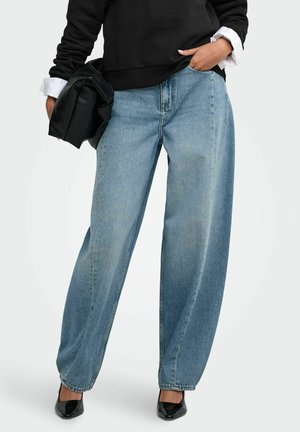 Personne portant un jean bleu clair à jambes larges, des chaussures noires pointues, un pull noir avec des poignets blancs, tenant un sac noir.