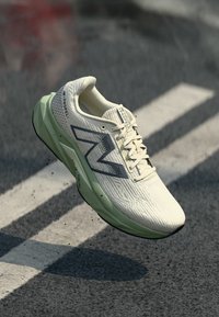 New Balance Laufschuh mit einem hellcremefarbenen Obermaterial mit strukturiertem Muster, grauen Akzenten und einer grünen Dämpfungsmittelsohle.