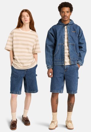 Kobieta i mężczyzna stoją obok siebie, ubrani w beżowe koszule w paski, dżinsowe szorty i mokasyny; mężczyzna ma również na sobie dżinsową kurtkę i tatuaże na nogach.