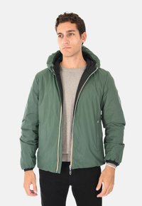 Uomo che indossa una giacca verde con cappuccio e zip sopra un maglione grigio e pantaloni neri, in piedi contro uno sfondo neutro.