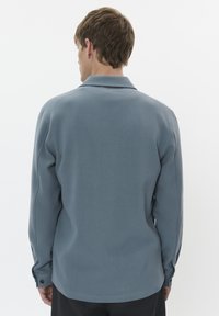 O persoană cu părul scurt stă cu spatele, purtând o cămașă cu mânecă lungă de culoare albastră deschisă, cu guler, pe un fond alb simplu.