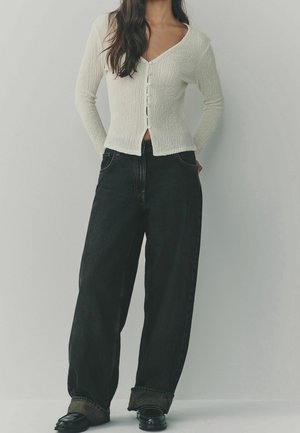 Femme portant un cardigan blanc boutonné et un jean noir large avec des chaussures noires, debout devant un fond gris clair uni.