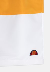 Kratšie nohavice s farebnými blokmi, pričom horná časť je jasne oranžová a dolná časť biela, s malým vyšitým logom na leme.