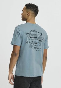 Lyseblå bomulds T-shirt med sorte grafik på ryggen, der viser danske madvarer og tekst: "Danish Foodie CLUB" og "Copenhagen EST 93."