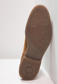 Hnědá semišová podešev s kruhovým vzorem na trati. Obsahuje embosovaný logo a text. Design zahrnuje flexibilní gumový materiál pro pohodlí.