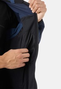 Handen die een binnenzak met een kleine ritssluiting openen in de binnenkant van een marineblauwe en zwarte jas.