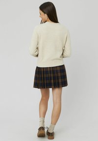 Un pull en tricot beige sur une jupe plissée à carreaux avec des tons marine, brun et doré. Des chaussures brunes avec des chaussettes beiges complètent l'ensemble.