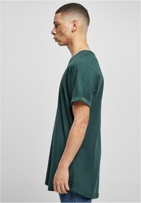 Camiseta de algodón verde con mangas cortas enrolladas, corte holgado y escote redondo, presentada de lado sobre un fondo neutro.