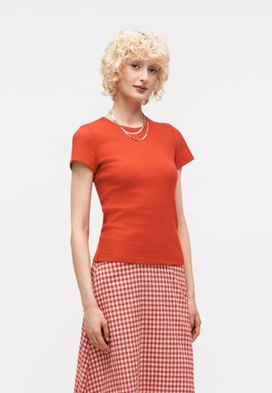 Lyshåret kvinde med kort krøllet hår bærer en tætsiddende rød kortærmet top, rød- og hvidternet nederdel og lagdelte perlekæder.