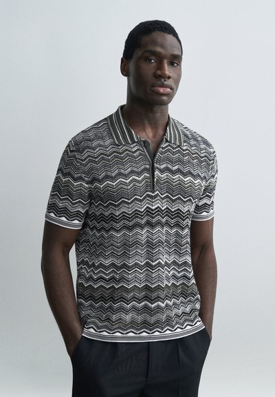 Homme debout les mains dans les poches, portant un polo à manches courtes noir et blanc avec un motif en zigzag et un pantalon foncé.