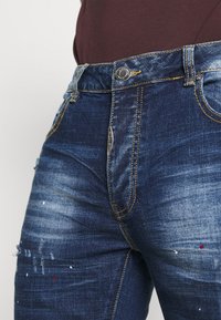 Tummansinisiä farkkuja, joissa on vaaleita kulumia, kuluneita yksityiskohtia ja hienovaraisia maalisuihkeita. Varustettu tavanomaisilla taskuilla ja metallisilla yksityiskohdilla.