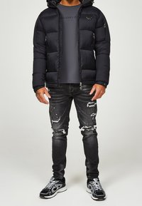 Mand iført en sort pufferjakke over en mørk t-shirt, slidte sorte jeans og sorte og hvide sneakers, står mod en ensfarvet baggrund.