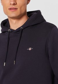 Marineblå hoodie lavet af bomuldsblandingsstof, med en front kangaroo-lomme, snoretræk i hætten, og et lille broderet logo på brystet.