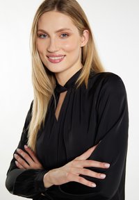 Schwarze Bluse aus glattem Stoff, mit hohem Ausschnitt, langen Ärmeln und einem Schlüsselaugen-Detail an der Front. Das Model hat die Arme verschränkt.