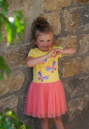 Junges Mädchen in gelbem und pinkem Kleid steht an einer Steinmauer, formt mit den Händen ein Herz und lächelt sanft.