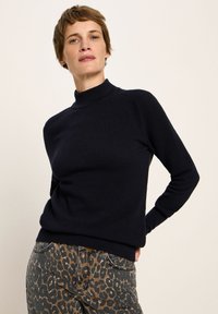 Frau mit kurzen Haaren, die einen schwarzen Langarmpullover und braune Leopardenmuster-Hosen trägt, steht vor einem schlichten hellen Hintergrund.