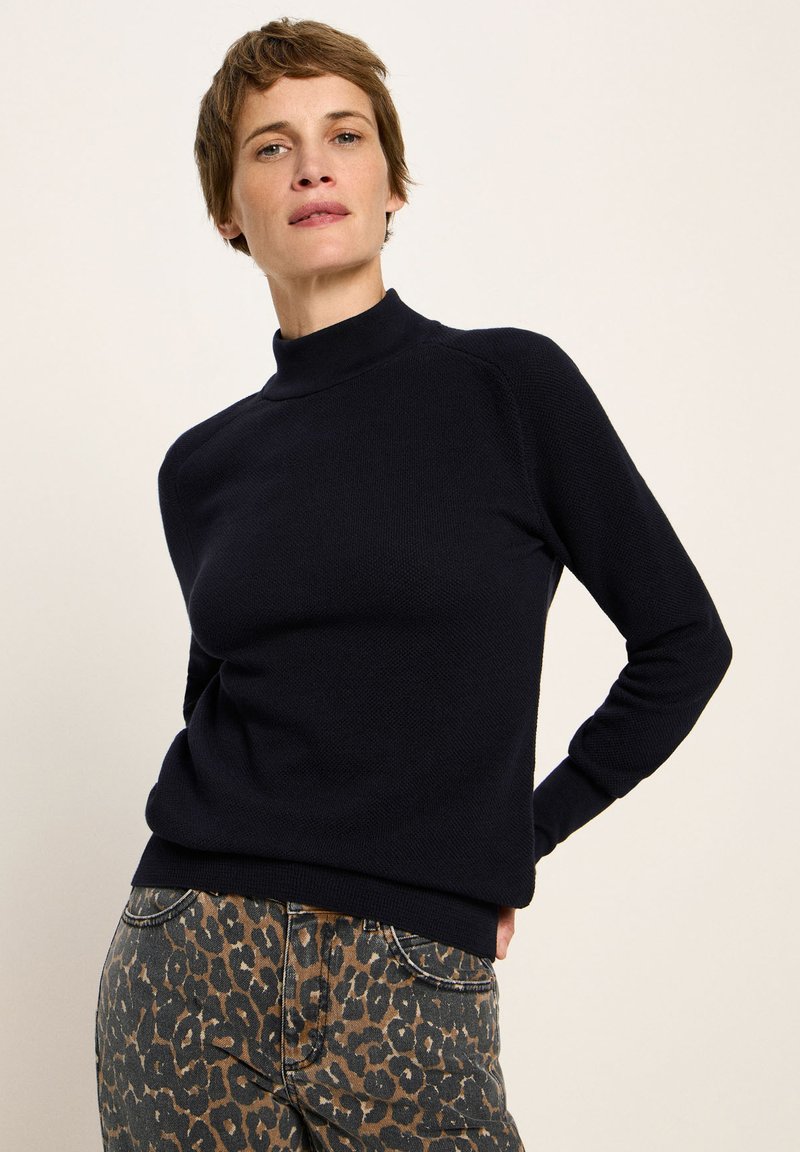 Frau mit kurzen Haaren, die einen schwarzen Langarmpullover und braune Leopardenmuster-Hosen trägt, steht vor einem schlichten hellen Hintergrund.