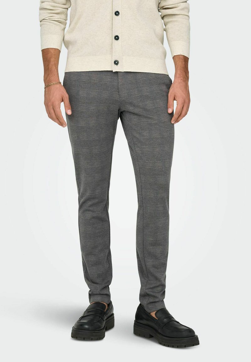 Homme portant un pantalon gris coupe slim, des mocassins noirs et un cardigan crème avec des boutons noirs, debout sur un fond clair uni.
