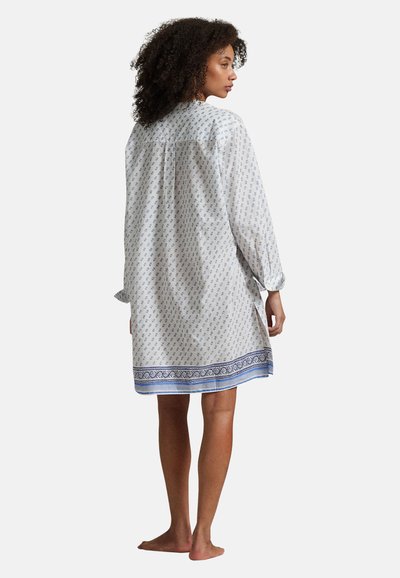 Femme aux cheveux bouclés, pieds nus, portant une robe blanche ample à manches longues avec un motif floral bleu et une bordure décorative bleue à l'ourlet.