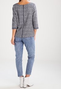 Navy- und weißes Strukturoberteil mit Rundhalsausschnitt, drei Viertelärmeln und einem Reißverschluss hinten, kombiniert mit hellblauen Jeans und weißen Ankle Boots.