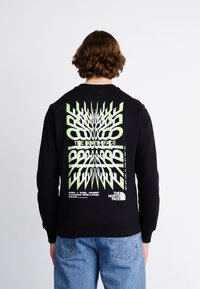 Persona vestida con un sudadera negra con el texto distorsionado en verde y blanco que dice "EXPLORE" y el logo de The North Face en la parte posterior, combinada con jeans azules.