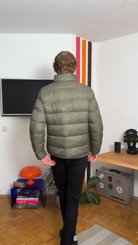 Maslinasto zeleni puffer jakna s visokim ovratnikom, kvadratnim uzorkom i rastezljivim manžetama, u kombinaciji s crnim hlačama. Pogled s leđa, nosi se unutra.