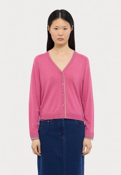 Cardigan rosa com decote em V, mangas longas e fecho de botão. Apresenta punhos e barra canelados com detalhes contrastantes. Usado sobre uma saia de ganga.