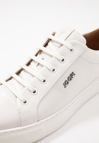 White leather sneaker with a textured surface, round toe, flat laces, metal eyelets, and a logo accent on the side.