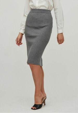 Kvinde iført en grå, ribstrikket pencilskirt med høj talje, en hvid langærmet skjorte og sorte åbne høje hæle.