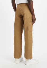Pantalones cargo caqui con un diseño de pierna recta, que cuentan con múltiples bolsillos, costuras visibles y un ajuste regular, combinados con zapatillas blancas.