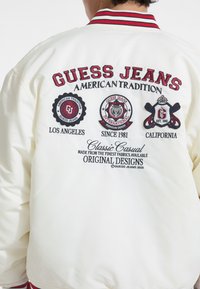 Weißer Bomberjacke mit gestickten Logos in Rot und Schwarz. Rippkragen in Rot und Schwarz. Enthält den Text "GUESS JEANS AMERICAN TRADITION."
