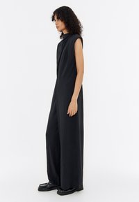 Black sleeveless jumpsuit with a high neckline, wide legs, textured fabric, and subtle detailing. Model wears black platform shoes.
