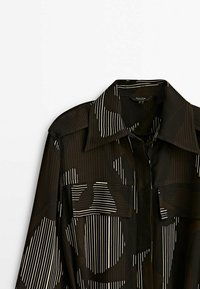 La camicia nera presenta motivi astratti con linee verticali bianche. È progettata con un colletto a punta e due tasche anteriori. Tessuto leggermente testurizzato.