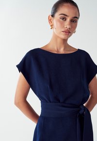 Mornarice plava haljina s kratkim rukavima, s brodskim izrezom i detaljem s vezanjem struka. Glatka tkanina s ležernim krojem, minimalistički dizajn.