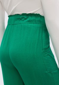 Πράσινο, υφασμάτινο παντελόνι με υφή, διαθέτει βολάν στη μέση και πλαϊνή τσέπη. Το ύφασμα φαίνεται ελαφρύ και αέρινο, κατάλληλο για καθημερινή χρήση.
