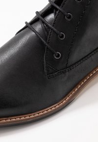 Bottillon en cuir noir avec bout rond, cinq œillets en métal, lacets noirs et une semelle extérieure marron contrastante avec des détails de couture.