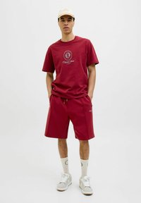 T-shirt en coton bordeaux avec design de logo, accompagnée d'un short bordeaux assorti. Le modèle porte une casquette crème, des chaussettes blanches et des baskets de couleur claire.