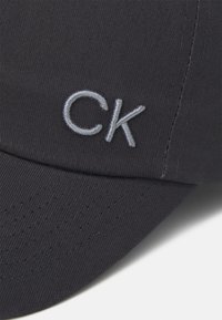 Tummanlaineinen baseball-lippis, joka on valmistettu teksturoidusta kankaasta ja jossa on hopean värinen brodeerattu "CK"-logo edessä sekä valmiiksi kaareva lippa.